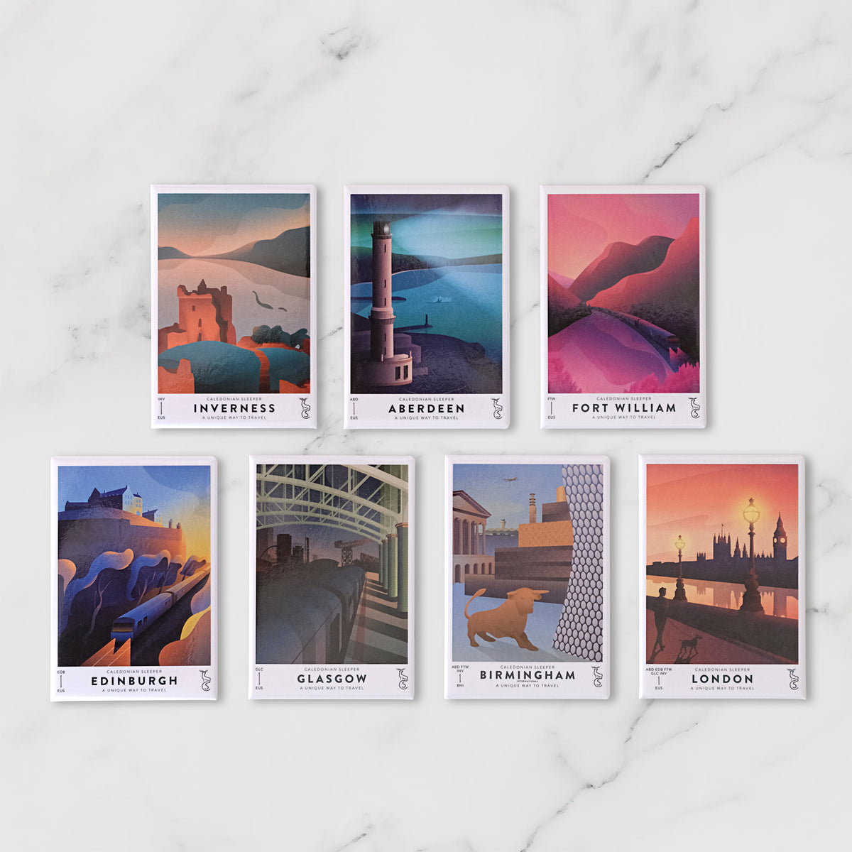 Seven illustrated travel Caledonian Sleeper Fridge Magnets featuring Edinburgh, Glasgow, Birmingham, London, Aberdeen, and Inverness, arranged on a white marble surface.